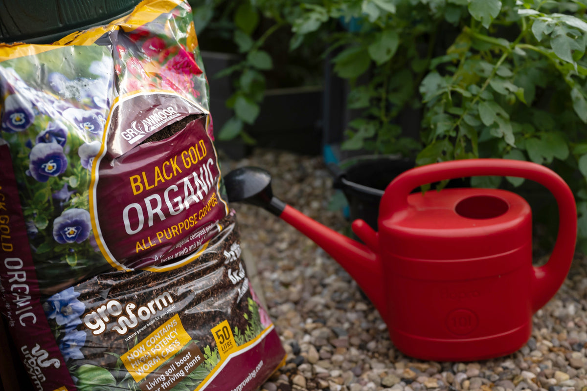 A bag of Gro Worm Black Gold Organic compost beside a red watering can in a garden